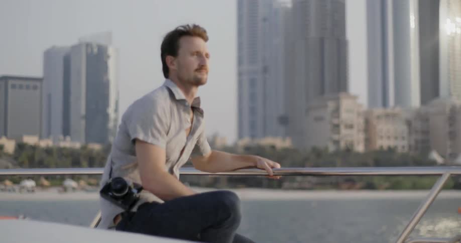 A man with a camera sits on a boat in the harbor of a city