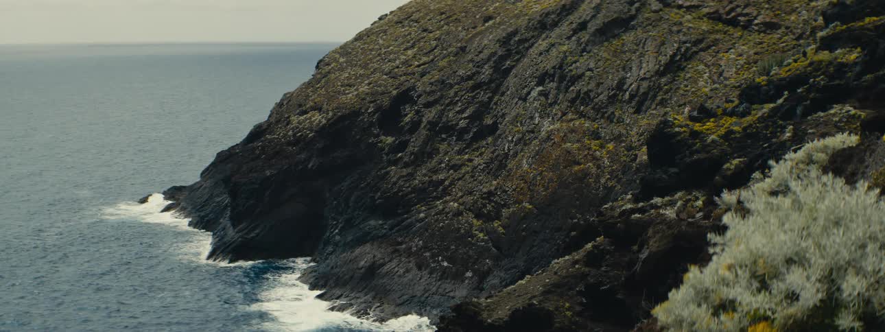 A static shot of ocean waves gently crashing against a rugged, moss-covered rocky shoreline