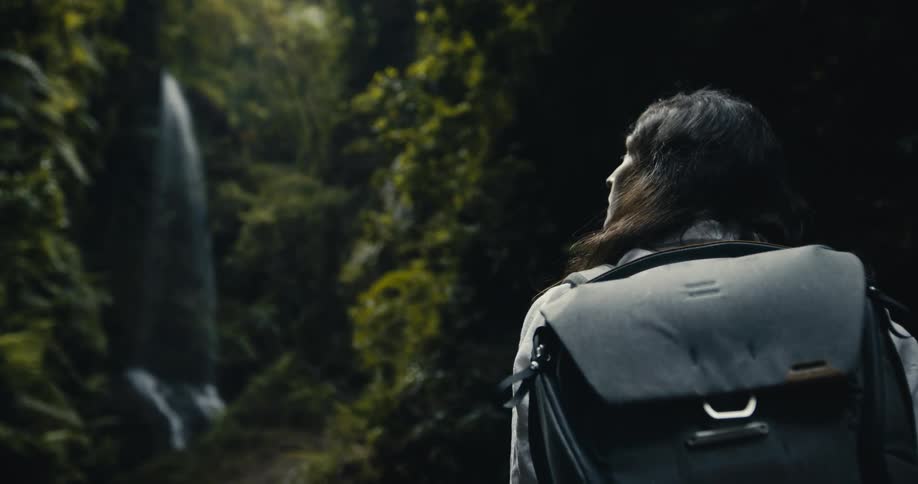 A solo hiker with a backpack pauses to gaze at a breathtaking waterfall cascading through a dense, lush jungle