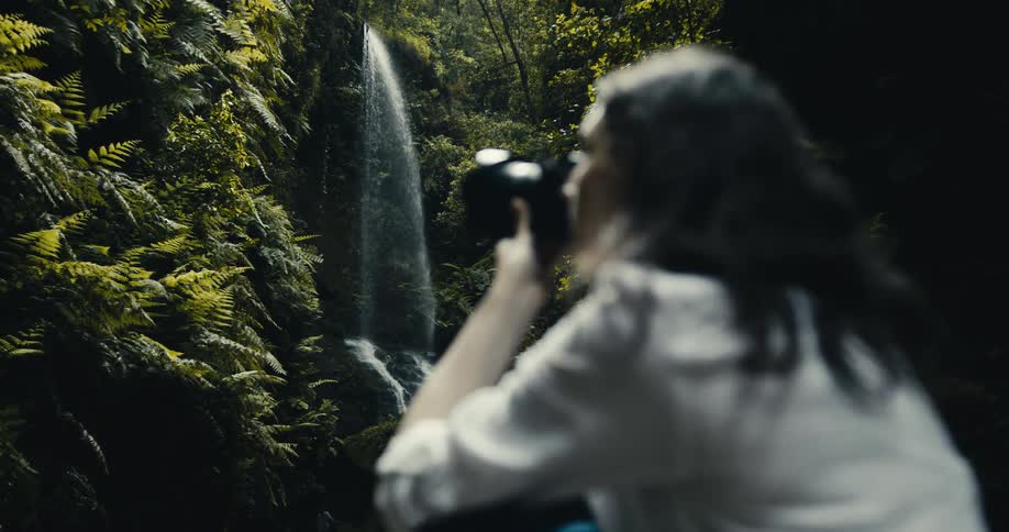 Over-the-shoulder shot of a woman photographing a beautiful waterfall deep within a lush, green forest