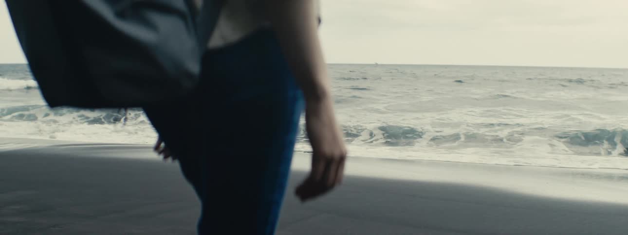 A person in blue jeans stands on a black sand beach as gentle ocean waves break on the shore in slow motion