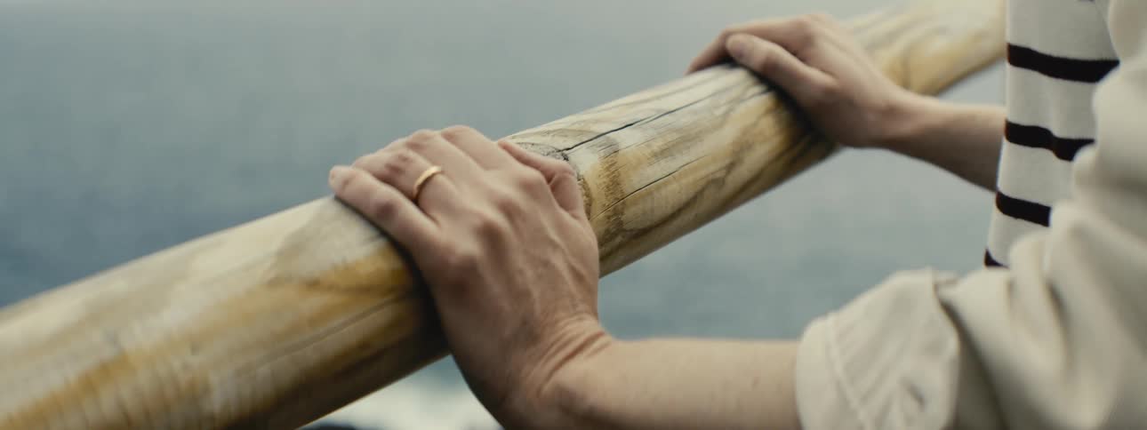 A contemplative person's hands with a wedding ring rest on a wooden railing overlooking the calm sea