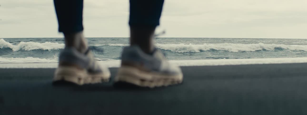 A person stands on a dark sand beach watching the ocean waves roll in on an overcast day, with their sneakers blurred in the foreground