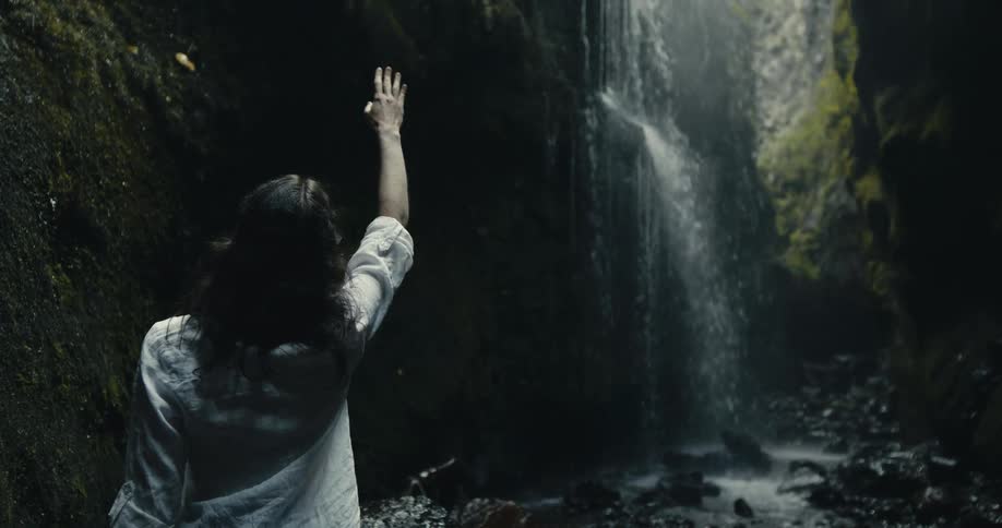 A mysterious woman slowly reaches for a mossy canyon wall, standing in awe before a cascading waterfall in a dark, serene grotto