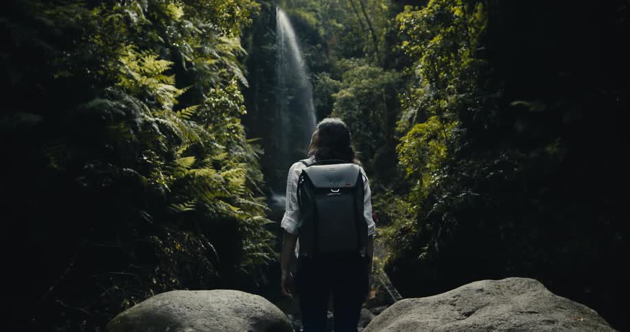 A lone adventurer with a backpack takes a moment to admire a breathtaking waterfall deep within a lush, green jungle