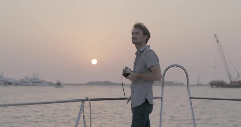 A man standing on a boat takes a photo at sunset