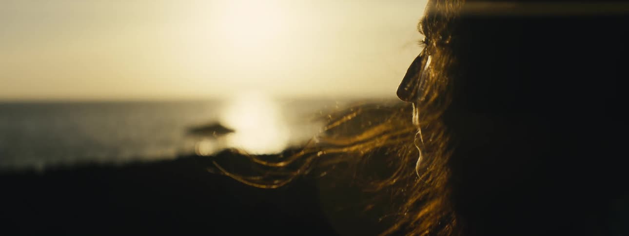 A woman watches the ocean sunset in slow motion as the wind blows through her hair