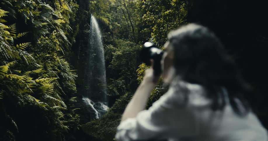 A photographer, out of focus in the foreground, captures a scenic waterfall cascading down rocks in a lush, green jungle