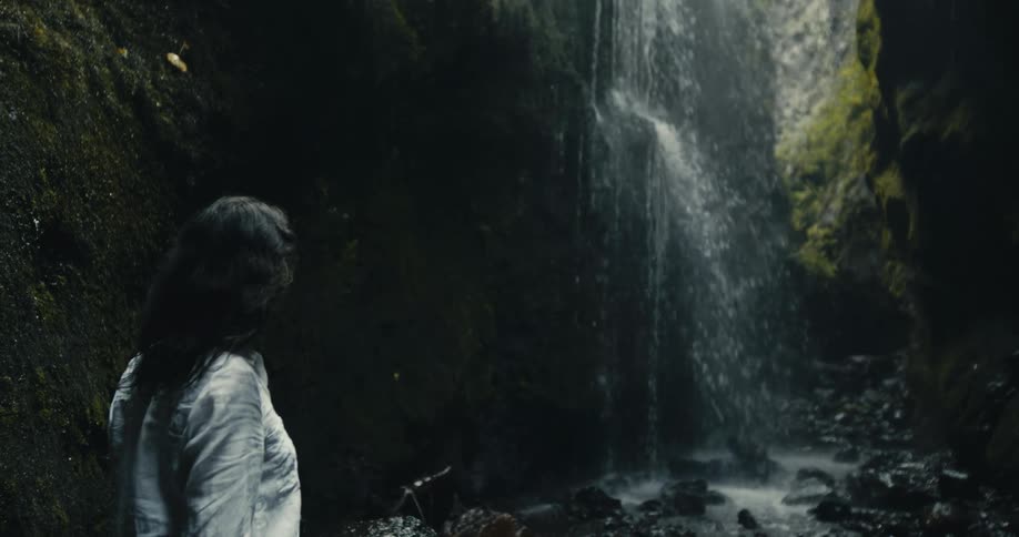A woman discovers a hidden waterfall in a dark, mossy canyon, illuminated by a single ray of light
