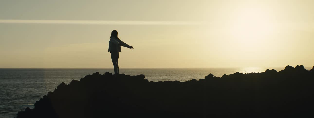 A lone woman silhouetted against a golden sunset slowly raises and lowers her arms while standing on a rocky coast overlooking the ocean