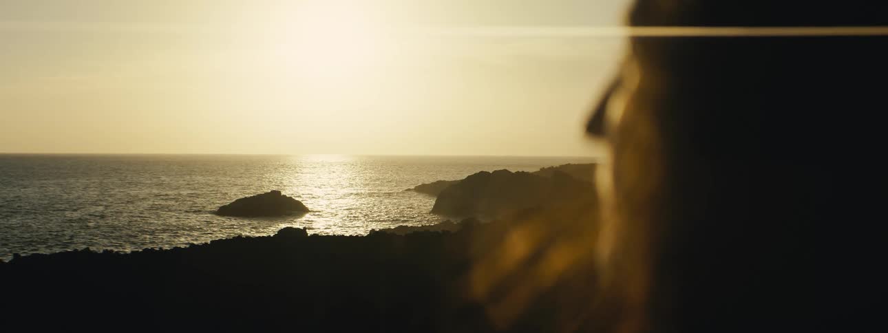 An over-the-shoulder view shows a person watching the golden sun reflect on the ocean along a rocky coastline