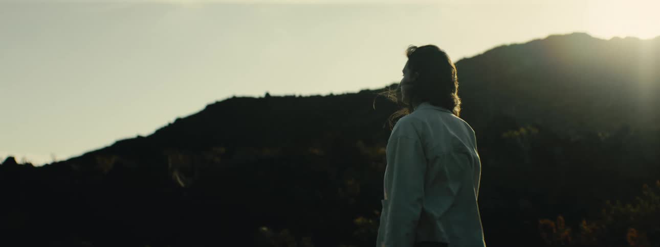 A woman stands thoughtfully on a hillside, her hair blowing in the wind as she gazes at the mountains during a beautiful golden hour