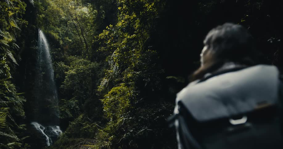 An over-the-shoulder shot of a person with a backpack admiring a beautiful waterfall hidden deep within a dark, lush rainforest