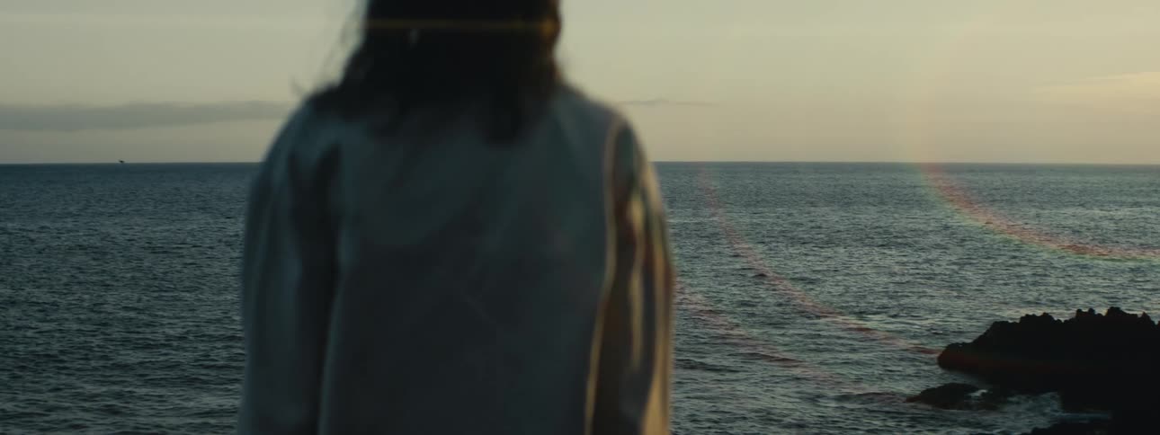 A solitary person stands on a rocky shore, peacefully contemplating the vast ocean at sunset