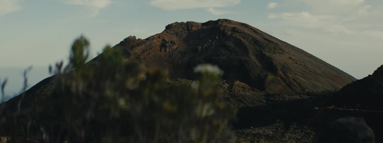 A rugged volcanic peak stands against a hazy sky, with out-of-focus desert flora in the foreground