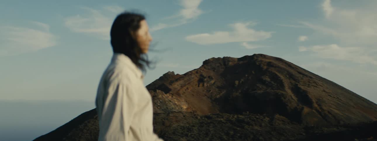 A woman with her back to the camera looks out at a large volcanic mountain at golden hour