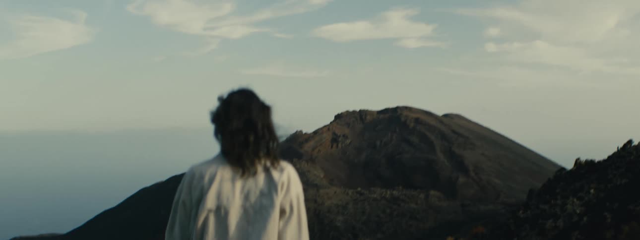 Slow motion shot from behind a person with windblown hair as they stand on a mountain looking out at a desolate volcanic landscape