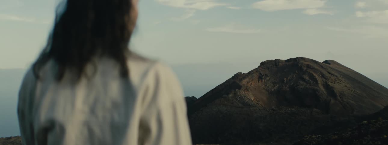A pensive person with windblown hair looks out at a vast volcanic landscape as the camera focus shifts to the distant mountain