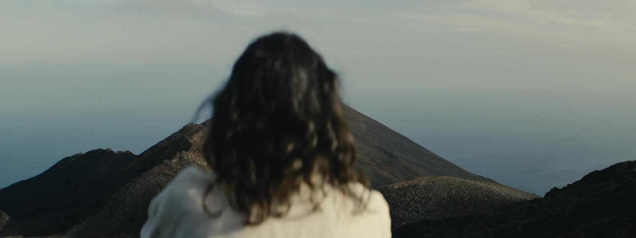 Over the shoulder shot of a woman with dark hair looking out at a large, barren volcano on a hazy day