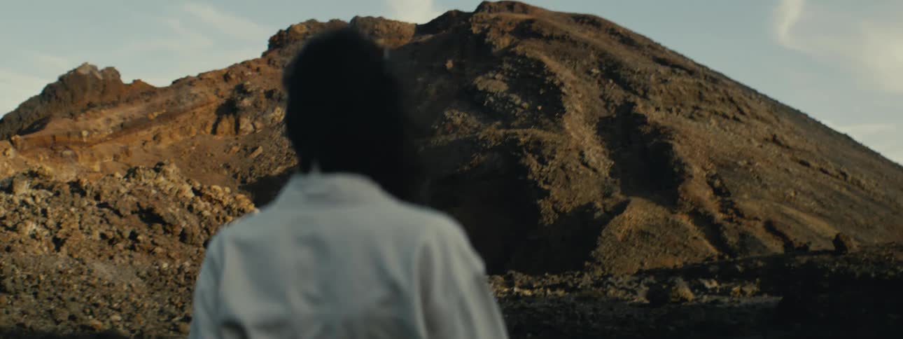 From behind, a person with windblown hair stands in a rocky, volcanic landscape, looking towards a large mountain