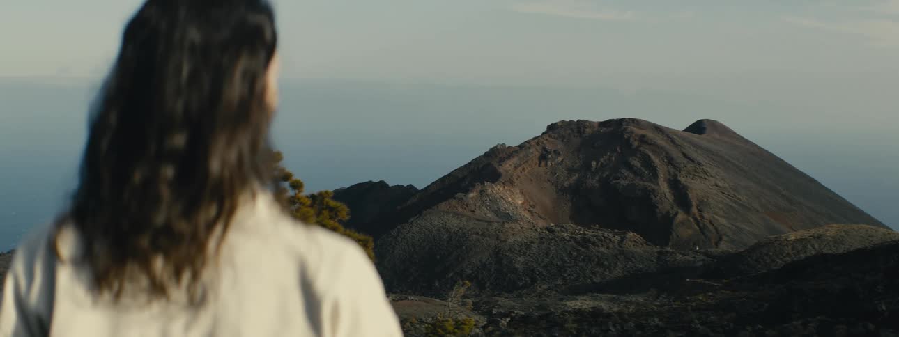 An over-the-shoulder shot reveals a person with long dark hair looking out at a majestic dormant volcano and a vast rocky landscape
