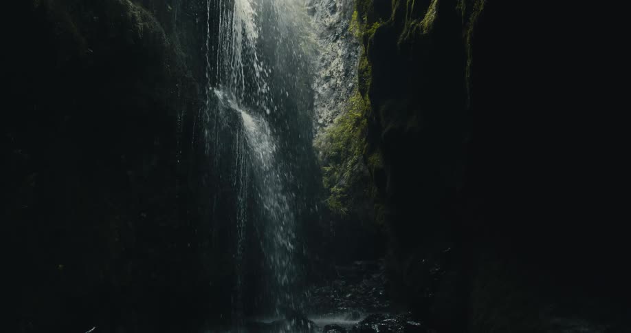 A hidden waterfall cascades down a mossy rock face into a dark, rocky stream within a secluded canyon