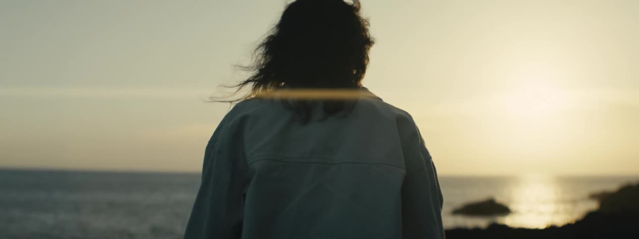 A woman with hair blowing in the wind looks out at the ocean during a peaceful golden hour sunset