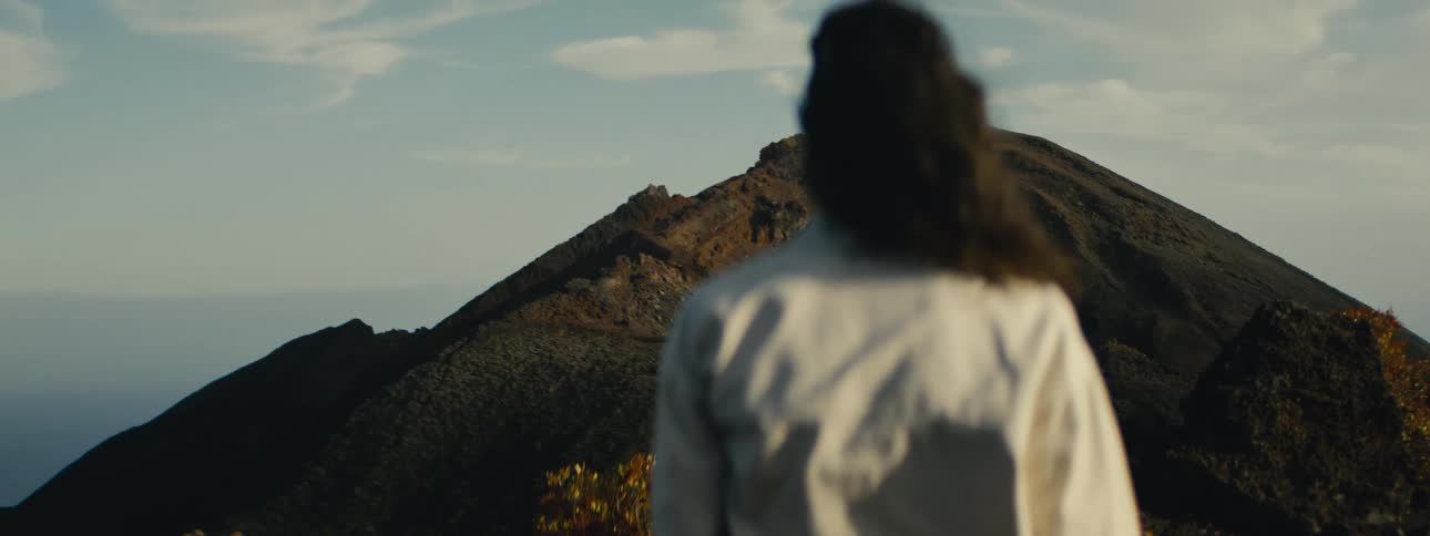 A person with long hair looks out at a vast volcanic landscape at golden hour