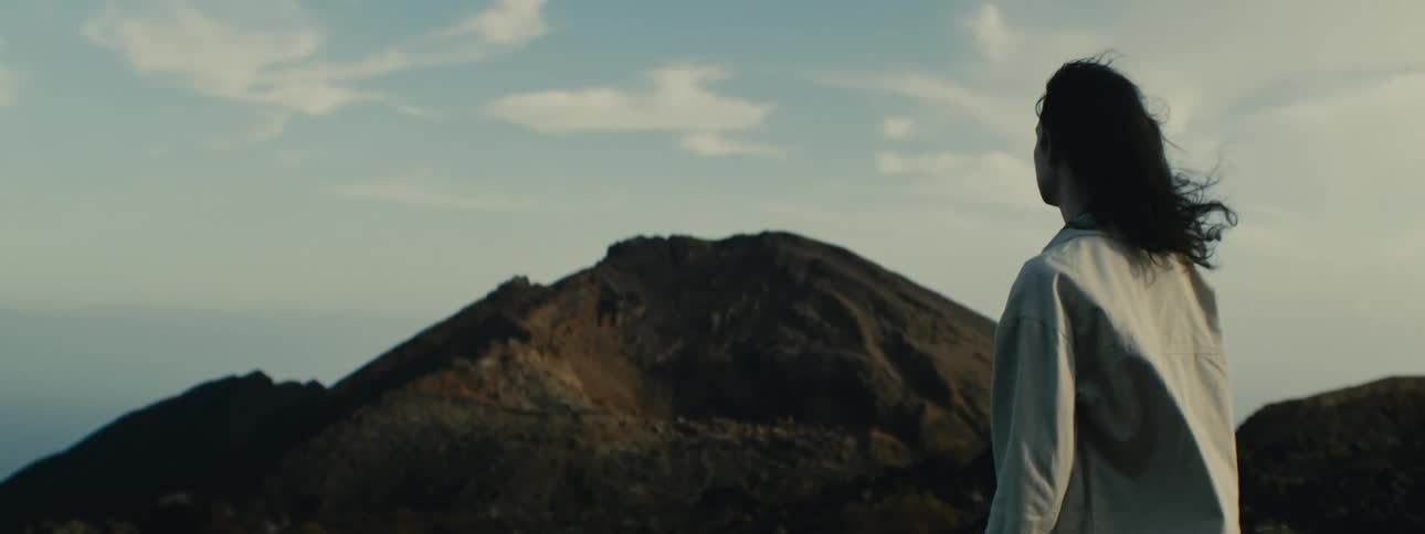 A person with long dark hair stands in a rocky, barren landscape looking out at a distant volcanic mountain
