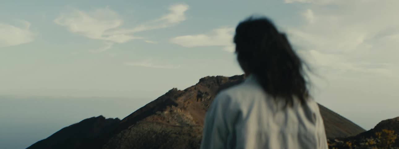 A lone figure stands with their back to the camera, hair blowing in the wind, looking out at a large, barren volcanic crater under a hazy sky