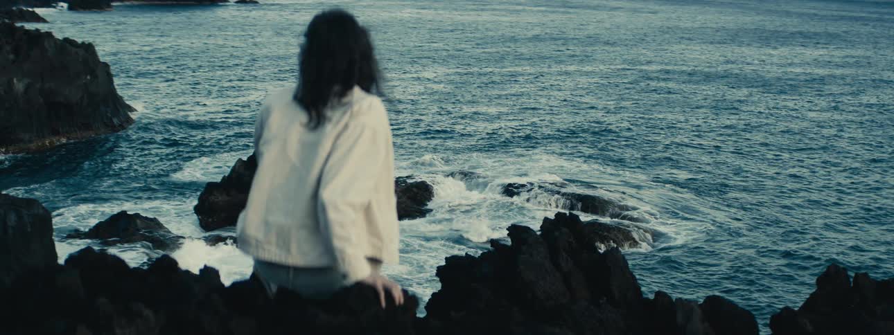 A solitary figure with dark hair sits on a rocky shoreline, gazing out at the vast ocean as waves crash against the volcanic rocks