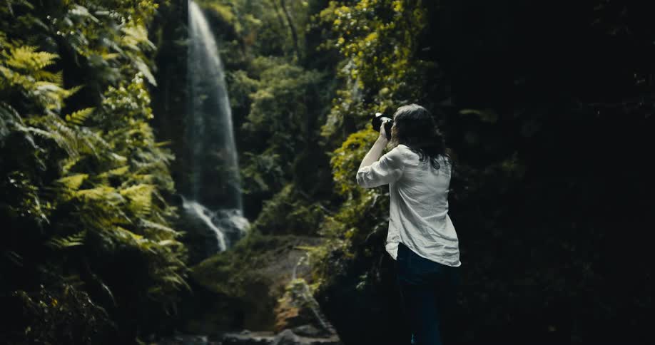 Slow motion shot of a female photographer capturing the serene beauty of a cascading waterfall in a dense, green jungle