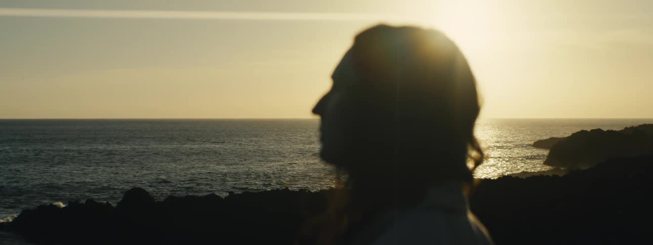 A silhouetted and out of focus person gazes at the golden sunset reflecting on the ocean from a rocky shore