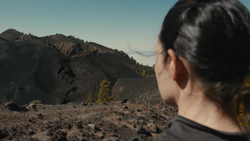 A woman pauses while hiking to gaze out over a vast, desolate volcanic mountain range that rises above a sea of clouds