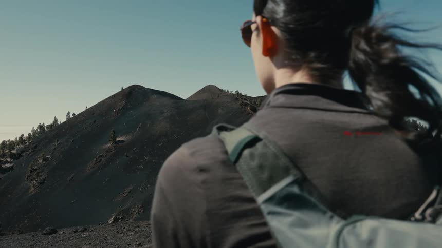 Slow motion over-the-shoulder shot of a female hiker with a backpack admiring the stunning volcanic mountain landscape on a sunny day