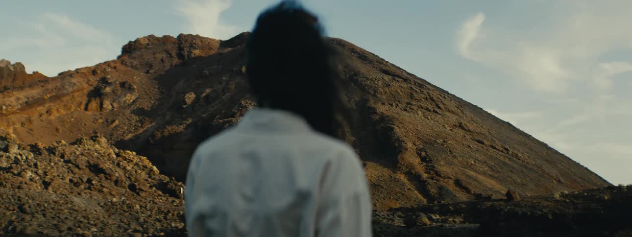 Over-the-shoulder shot of a person with hair blowing in the wind, looking out at a vast, volcanic mountain landscape