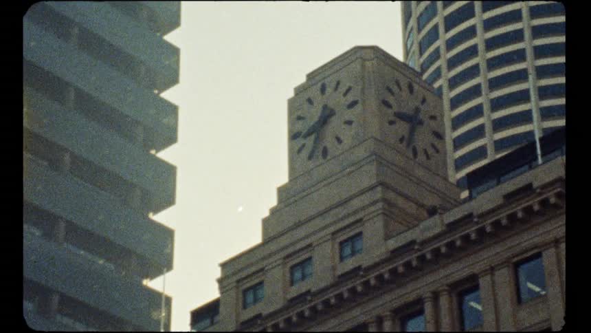 Authentic vintage footage shows a historic clock tower dwarfed by modern architectural giants in a downtown city setting