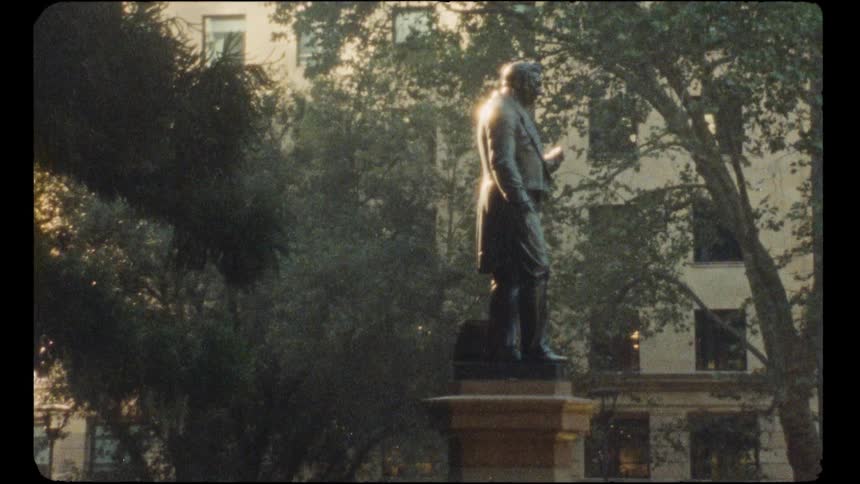 A vintage shot captures a bronze statue of a historical figure in a city park during the golden hour