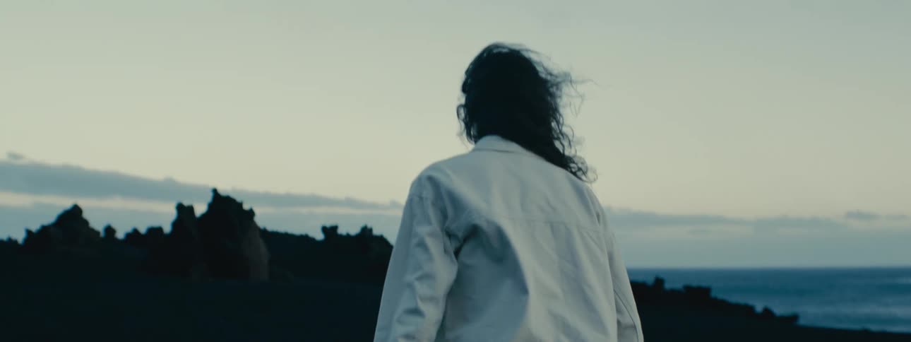 Slow motion shot from behind of a person with windblown hair walking through a dramatic black sand volcanic landscape towards the distant sea