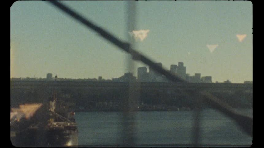 Vintage Super 8 film captures a hazy city skyline and a cargo ship on a river, filmed from a moving vehicle crossing a bridge