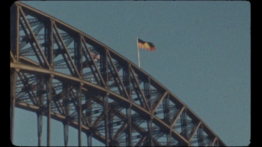 A vintage Super 8 film shot of the Australian Aboriginal Flag flying proudly atop the Sydney Harbour Bridge against a clear blue sky