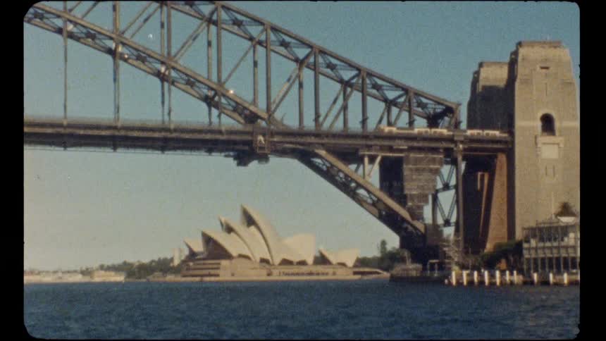 An archival shot captures trains crossing the iconic Sydney Harbour Bridge with the famous Sydney Opera House visible in the background on a sunny day