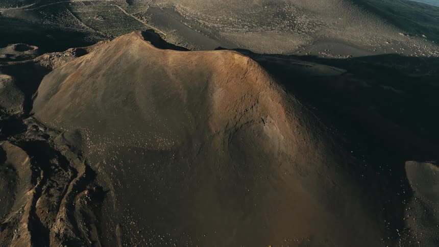 A dramatic aerial shot orbits the dark, ashen slopes and reddish crater of a massive dormant volcano