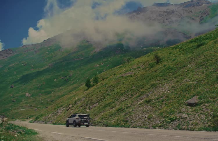 An SUV with a bicycle on the back drives up a winding mountain pass through a lush green valley on a beautiful day