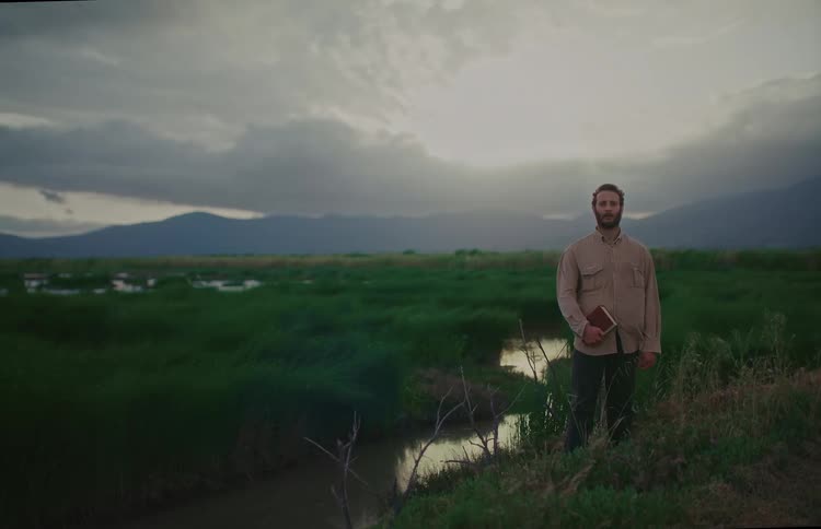 A pensive man holding a book stands in a windswept marsh at dusk with mountains in the background