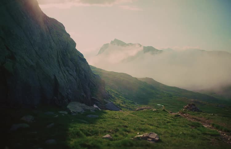 A serene view of a rocky cliff overlooking a green mountain valley with clouds rolling over distant peaks at sunrise