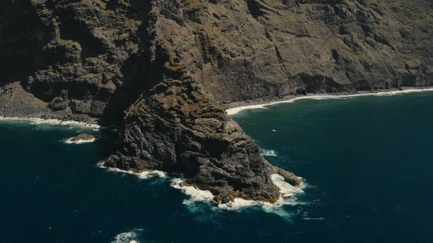 A static aerial view captures the dramatic, rocky coastline where massive cliffs meet the deep blue ocean