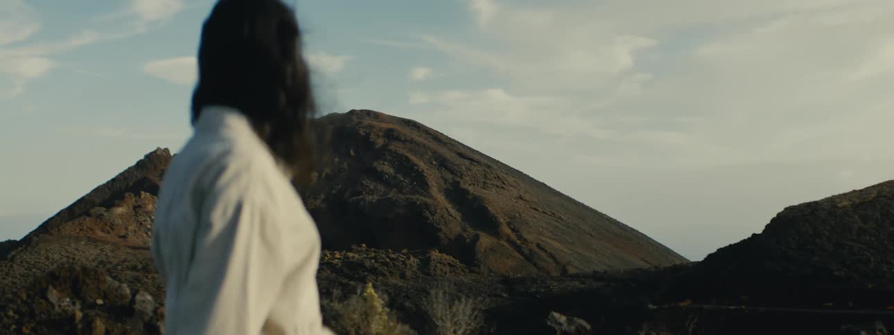 A person with long dark hair stands in a vast, windy volcanic landscape and gazes at a distant mountain