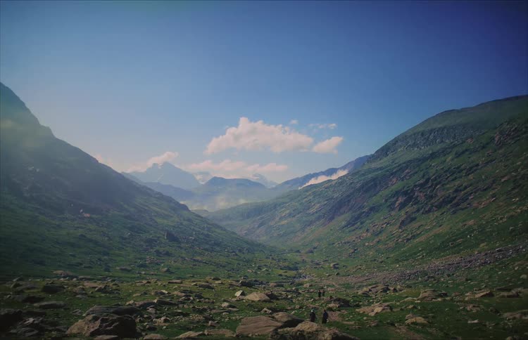 An epic wide shot of a few distant hikers traversing a vast, lush green mountain valley under a clear blue sky