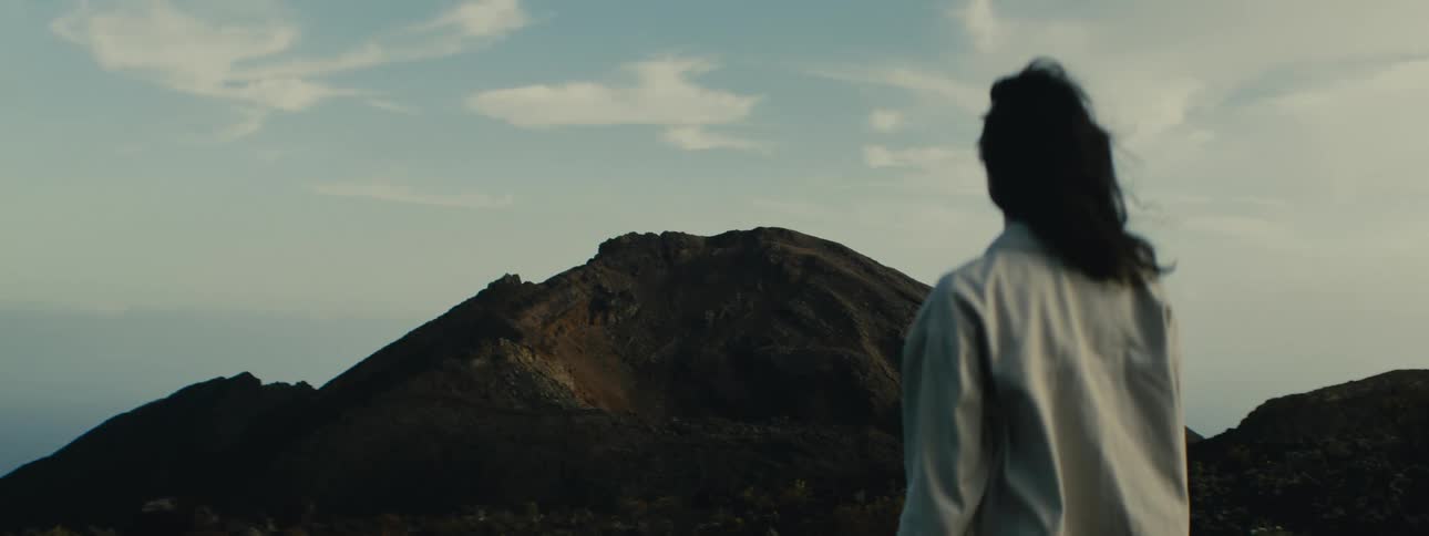 From over the shoulder, a person with windblown hair gazes at a distant, dark volcanic mountain under a pale sky